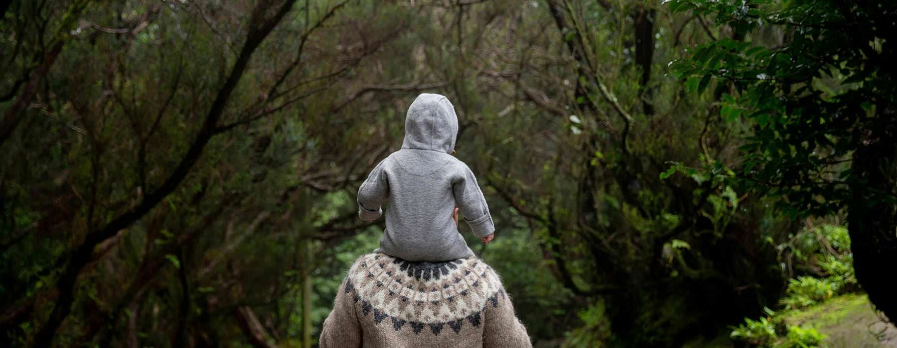 man holds his child on his shoulders while walking through a forest
