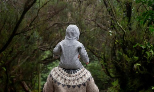 man holds his child on his shoulders while walking through a forest